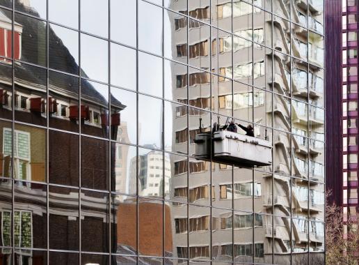 Hoge gebouwen gezien vanuit spiegelend flatgebouw met glazenwassers in bakje tegen gebouw aan.