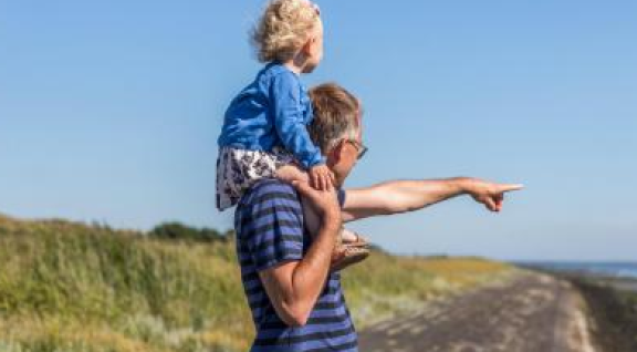 Een kind op de nek bij een man die wijst naar de verte, met op de achtergrond groen en een weg