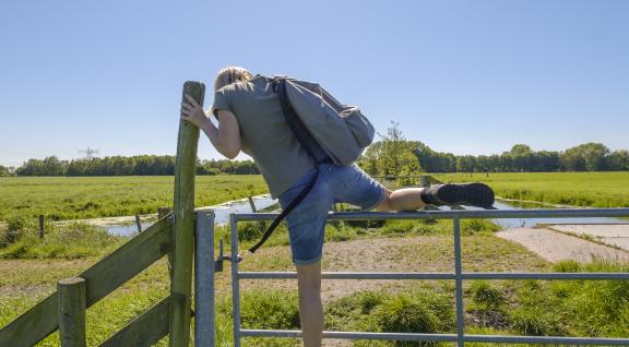 Een vrouw met wandelschoenen aan klimt over een hek in een weiland